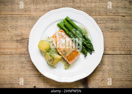 Vue de dessus du filet de saumon grillé avec des pommes de terre et des asperges garnies de sauce à l'aneth sur une assiette blanche et une ancienne table en bois Banque D'Images