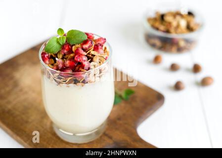 Petit-déjeuner sain concept. Crème fraîche vanille avec granola dans un verre garni de grenadier et menthe disposées sur une table en bois blanc. Banque D'Images