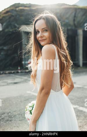 Portrait d'une mariée amoureuse regardant par-dessus son épaule debout sur la route de montagne pendant qu'elle est à la séance photo le jour de son mariage. Rustique Banque D'Images