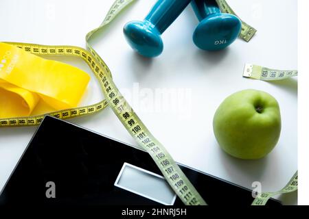 Une pomme, une balance, une bande élastique de forme physique et des haltères reposent sur un fond blanc, ils sont enchârés avec un ruban de mesure Banque D'Images