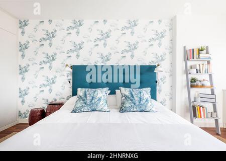 Tête de lit capitonnée en tissu bleu capitone avec coussins assortis à motifs floraux, papier décoratif sur le mur et une étagère avec un livre en un c Banque D'Images