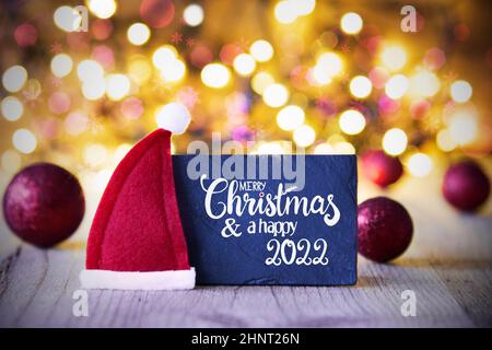 Sighn Wth English Calligraphie Joyeux Noël et heureux 2022.Décoration boule de Noël violette avec chapeau de père Noël rouge.Arrière-plan de lumières étincelantes Banque D'Images