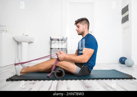 Jeune homme utilisant la ceinture de yoga tout en faisant de l'exercice sur le tapis de fitness à la clinique de réadaptation. Banque D'Images