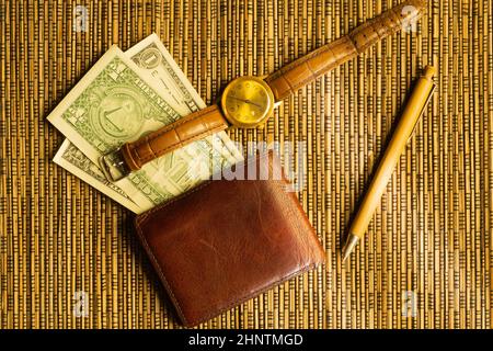 Des dollars dans un portefeuille avec une montre-bracelet et un stylo en or. De l'argent sur un fond de bambou en bois. Fond financier Banque D'Images