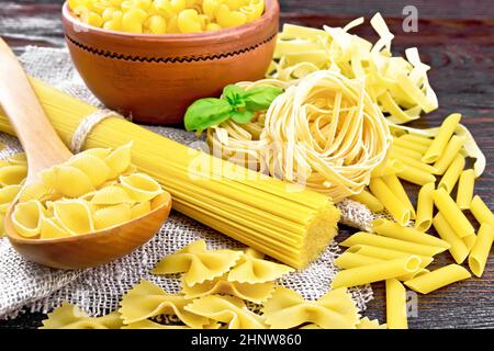 Pâtes de coquillages dans une cuillère, tuyau de rigate dans un bol, spaghetti, tagliatelle, penne, farfalle, trenette sur serviette de toile de jute sur fond de panneau en bois foncé Banque D'Images