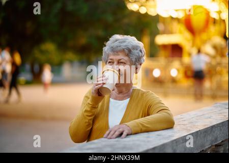 Jolie boisson granny café dans le parc d'attractions d'été, éclairage lumineux sur fond. Style de vie des personnes âgées. Grand-mère drôle ayant du plaisir à l'extérieur, Banque D'Images