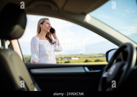 Jolie femme d'âge moyen au volant de sa voiture, ayant une pause sur un long voyage, faisant un appel téléphonique Banque D'Images