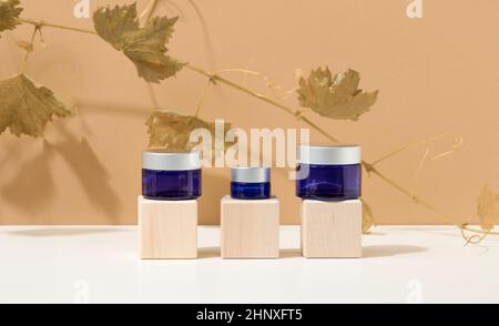 produits cosmétiques dans un pot en verre bleu avec un couvercle gris sur un podium en bois en cubes, derrière une branche de raisins avec des feuilles vertes. Vide pour le branding Banque D'Images