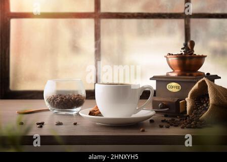 Tasse de café chaud avec sac plein de grains et moulin manuel sur table en bois et rebord de fenêtre. Vue avant. Composition horizontale. Banque D'Images