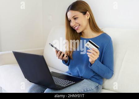Femme brésilienne tenant une carte de crédit et un smartphone utilisant un ordinateur portable pour faire des achats en ligne à la maison. OTP, authentification à deux facteurs, pix et jeton pour Banque D'Images