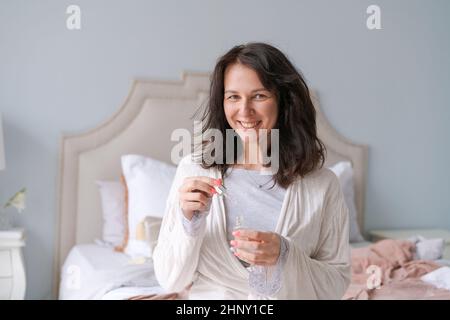 Concept beauté et soins de la peau. Le visage de la femme applique du sérum pour le visage de l'arbre à thé. Belle fille brune naturelle sans maquillage applique du sérum sur son visage avec une pipette. Dans un peignoir dans la chambre sur le lit Banque D'Images