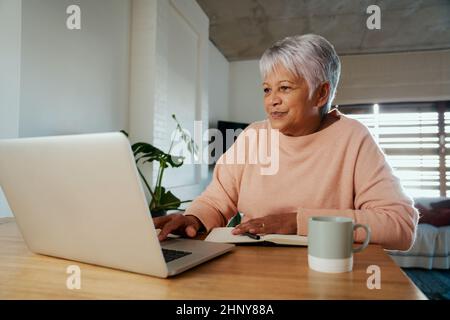 Une femme âgée, multiethnique, fait défiler son ordinateur portable tout en prenant des notes dans son ordinateur portable dans sa maison moderne. Banque D'Images