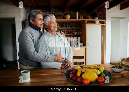 Heureusement, un couple biracial âgé à la retraite se tenant debout, embrassant et regardant au loin. Un style de vie sain dans une cuisine moderne. Banque D'Images