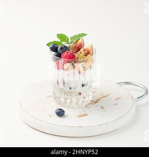 verre transparent avec granola versé avec yaourt, sur les framboises mûres, les bleuets et les figues sur une table blanche. Petit déjeuner sain Banque D'Images