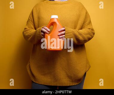 femme dans un pull tient une bouteille en plastique orange avec du gel de lavage liquide.Linge et corvées, fond jaune Banque D'Images