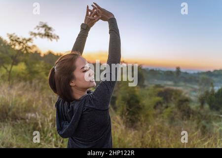 Belle jeune femme asiatique en veste d'échauffement qui étire ses bras pendant sa pause après son entraînement matinal dans un parc, lever du soleil bleu clair Banque D'Images