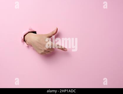la main femelle dépasse d'un trou déchiré sur fond de papier rose, une partie du corps pointe avec l'index sur le côté Banque D'Images