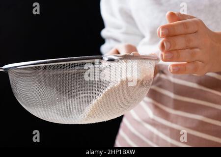 Une femme en gros filtre la farine de blé à travers un tamis dans un bol de mélange pour la préparation de la pâte pour le gâteau et la boulangerie sur fond noir. Pétrissez la pâte pour obtenir une pâtisserie maison. Banque D'Images