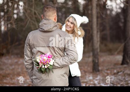 Joyeux sourire jeune couple dans les vêtements décontractés de dessus stand de rencontre des yeux tandis que l'homme cacher le bouquet de fleurs blanches, roses. Banque D'Images