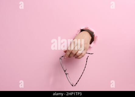 femme tenant des lunettes, partie du corps qui dépasse du trou déchiré sur fond de papier rose Banque D'Images