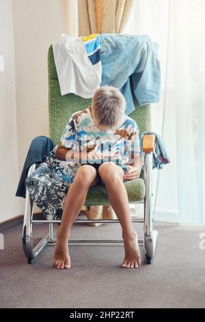 Un garçon blond est assis dans un fauteuil avec des piles de vêtements sales et joue à des jeux en ligne via une tablette. Un élève passe du temps libre à se détendre avec les technologies modernes Banque D'Images