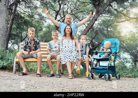 Bonne grande famille pose assis sur le banc dans le parc vert. Les parents souriants et les fils de différents âges aiment se reposer dans la nature contre les arbres et les buissons Banque D'Images