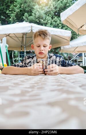 Un écolier aux cheveux équitables en chemise colorée joue assis à table en attendant la nourriture. L'enfant s'assoit dans l'ennui près des canopées contre les arbres verts de près Banque D'Images