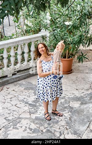 La jeune mère avec un bébé garçon aux bras se tient sur la terrasse du parc avec un large sourire heureux. La famille de maman et de fils profitent d'un temps tropical chaud Banque D'Images