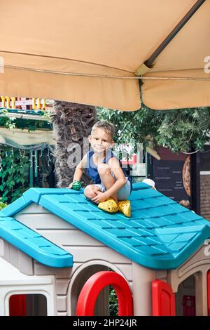 Le garçon d'avant-garde en vêtements d'été est assis sur le toit lumineux de la maison de jouets dans l'aire de jeux du restaurant. Un enfant aux cheveux équitables passe du temps libre à jouer à l'extérieur Banque D'Images
