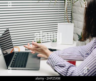 une femme dans une chemise rayée se trouve sur un bureau blanc et communique en ligne via un ordinateur portable.Appel vidéo, blogging.L'homme se gestuelle activement avec ses mains Banque D'Images