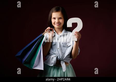 mignon petite fille tient beaucoup de sacs de shopping et point d'interrogation sur fond sombre. Banque D'Images