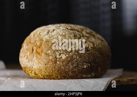 le pain rond avec une croûte croustillante vive repose sur du parchemin sur une table en bois, arrière-plan sombre, vue latérale Banque D'Images