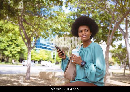 Femme d'affaires afro-américaine de taille moyenne qui regarde loin tout en étant assise avec un téléphone et une tasse jetable Banque D'Images