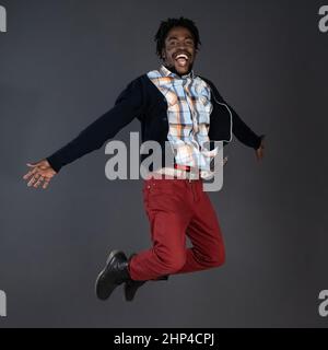 Émotionnellement motivé, positif, insouciant africain jeune homme américain en jeans rouges et chandail noir heureux sauter en studio isolé sur fond gris foncé. Banque D'Images