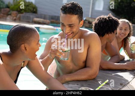 Un homme et une femme biracial souriants qui parlent entre eux avec des amis au bord de la piscine par beau temps Banque D'Images