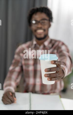 Accent sélectif sur la main d'homme afro-américain avec une tasse en carton ou une tasse jetable pour le café ou le thé. Gobelet blanc jetable avec fil en plastique bleu dans la main de l'homme. Concept de boîtes jetables. Banque D'Images