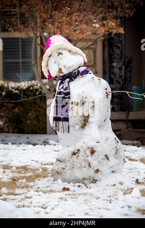 Bonhomme de neige avec chapeau rose et foulard violet et vieux cigare dans sa bouche debout devant la maison. Banque D'Images