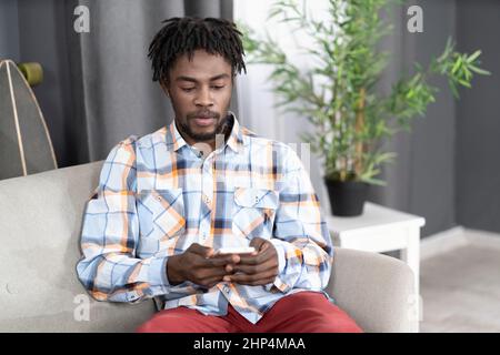 Un homme afro-américain concentré ou intéressé utilise un smartphone pour vérifier les médias sociaux ou mettre à jour les informations. Portrait d'un jeune homme tenant un smartphone assis sur le canapé. Concept de médias sociaux. Banque D'Images