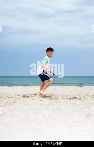 Le garçon s'entraîne sur le sable au bord de la mer. Entraînement sur les jambes. Préparation générale de l'allumage. Banque D'Images