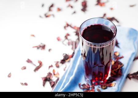 Thé Hibiscus dans un grand verre sur une serviette bleue. Thé à l'hibiscus sain et pétales d'hibiscus secs sur fond blanc. Copier l'espace Banque D'Images