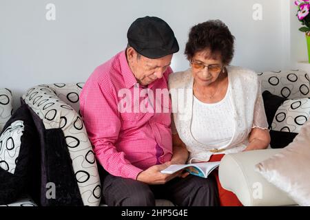 Couple âgé assis sur un canapé lisant un livre ensemble dans le confort de leur maison. Banque D'Images