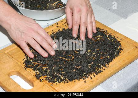 Les mains des femmes versent le mélange de thé sur une planche de bois pour sécher. Le processus de préparation d'un mélange de thé à partir de thé noir, de zeste de citron et d'orange, de morceaux de cin Banque D'Images