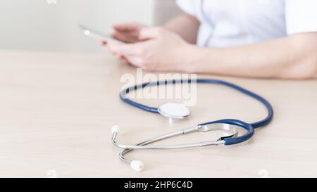 Une femme médecin méconnaissable s'assoit à un bureau et écrit un message sur un smartphone. Le praticien tient un téléphone dans ses mains et répond aux patients Banque D'Images