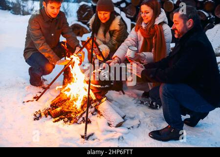 Groupe d'amis se rassemblant autour du feu de joie dans la cour arrière, boire du thé et réchauffer les mains. Deux couples heureux se détendant et profitant de la saison d'hiver tout en s'asseyant Banque D'Images