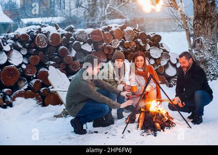 Groupe d'amis se rassemblant autour du feu de joie dans la cour arrière, boire du thé et réchauffer les mains. Deux couples heureux se détendant et profitant de la saison d'hiver tout en s'asseyant Banque D'Images