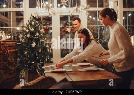 Deux jeunes femmes ad man mettant la table de Noël festive ensemble à la maison, la mère heureuse et la fille se préparant pour Noël célébration dans grand confortable rustique f Banque D'Images