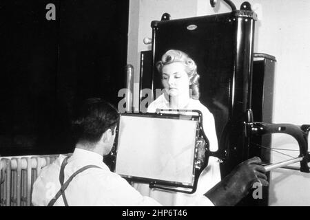 L'illustration montre un technicien masculin prenant un fluoroscope à rayons X d'une patiente. Le tube à rayons X se trouve derrière le patient et un écran en verre avec un revêtement fluorescent devant le patient rend l'image visible. Les examens par fluoroscope ont donné des expositions aux rayonnements beaucoup plus élevées que les images radiographiques sur film. Cette image a été utilisée pour démontrer le mythe de l'exposition au rayonnement pendant la procédure de rayons X. ca. Date de création: 1940 Banque D'Images