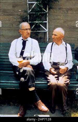 Deux mâles caucasiens âgés dans des manches de chemise assis sur un banc à l'extérieur et boivent de la bière ca. 1980 Banque D'Images