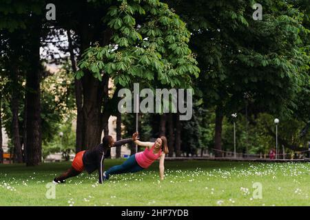 Deux jeunes femmes souriantes et diverses en vêtements d'entraînement athlétique font un exercice de planche dans le parc. Banque D'Images