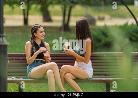 Photo à travers les buissons de parc de deux jeunes femmes joyeuses qui boivent leur smoothies en étant assis sur un banc à l'extérieur Banque D'Images